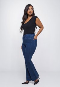 Donkerblauwe uitlopende jeans met vijf zakken, hoge taille en zichtbare stiksels. Zwart getailleerd top met een ronde hals. Puntige zwarte hakken.