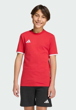 Ragazzo sorridente con capelli castani che indossa una maglietta rossa Adidas e pantaloncini neri Adidas su uno sfondo chiaro e uniforme.