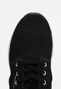 Partie avant d'une basket en tissu tricoté noir avec des lacets passés à travers des œillets métalliques, montrant un motif texturé détaillé sur la partie supérieure.