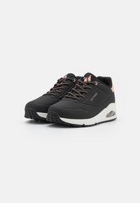 Scarpe sportive nere con superficie testurizzata, dotate di accenti bronzei, lacci e una suola intermedia bianca con un cuscinetto d'aria trasparente.
