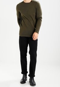 Maglietta verde oliva a maniche lunghe con scollatura rotonda, abbinata a pantaloni neri e stivaletti neri eleganti.