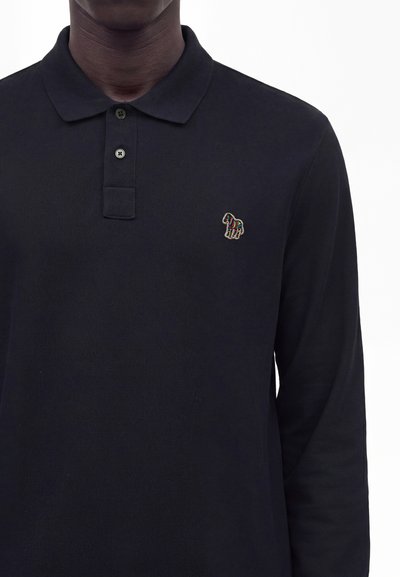 Homme portant un polo noir à manches longues avec deux boutons et un petit logo éléphant coloré sur le côté gauche de la poitrine.