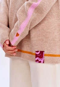 Beiger Pullover mit rosa und orangefarbenen Streifen. Verfügt über ein kleines Herzdetail am Ärmel und ein gebrandetes Etikett am Saum mit einem bunten Design.