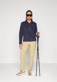 Person wearing sunglasses, navy fleece jacket, beige pants, and sneakers, holding two black hiking poles, standing against a plain background.