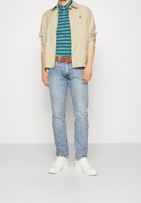 Lehká tanová bunda přes zelený a modrý pruhovaný polo, spárovaná se světle modrými džínami a b ílými teniskami, s hnědým pásem.