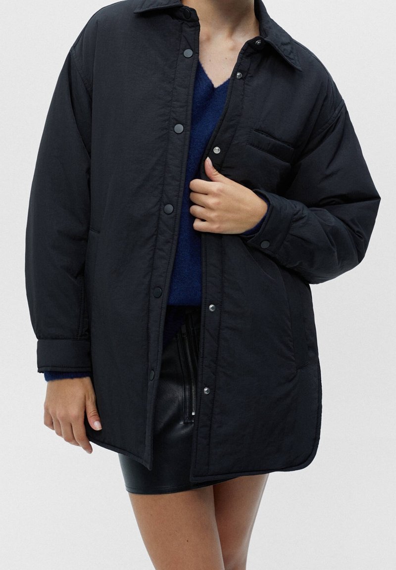 Femme portant une veste noire boutonnée par-dessus un pull bleu et une mini-jupe en cuir noire, debout devant un fond clair uni.