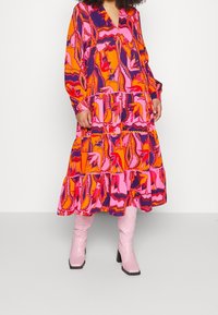 Lange, gelaagde jurk in een levendig oranje, roze en paarse bloemenpatroon. Gemaakt van lichte stof met lange mouwen en een V-hals.