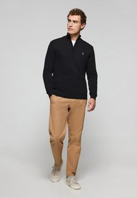 Svart zip-up tröja med ribbade muddar, bärs över en vit t-shirt, ihop med beige chinos och ljusa sneakers. En enkel logotyp på bröstet.