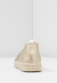 Zapato slip-on perforado beige con un acabado metálico suave, respaldo redondeado y suela de goma blanca con acentos ligeros texturizados.