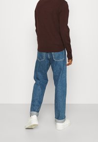 Eine Person trägt hochgekrempelte blaue Jeans, weiße Turnschuhe und einen langärmeligen dunkelbraunen Pullover, während sie vor einem schlichten weißen Hintergrund steht.