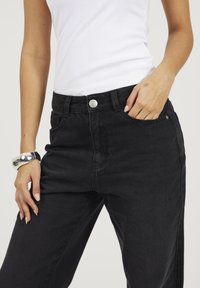 Jeans neri con una vestibilità comoda, caratterizzati da una chiusura a bottone, tasche frontali e una superficie in denim liscia. Abbinati a un top bianco senza maniche.