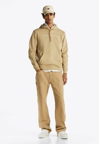 Beżowa bluza z kapturem z logo "TOMMY JEANS", kieszeń kangurka, połączona z pasującymi beżowymi spodniami cargo. Białe sneakersy dopełniają stylizację.