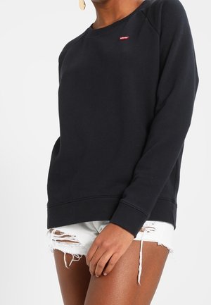 Henkilö pukeutunut mustaan pitkähihainen collegepaitaan, jossa on punainen logo, ja valkoisiin kuluneisiin farkkushortseihin, seisoo käsi reisillä lepäämässä.