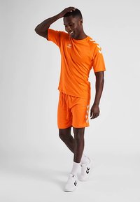Oranje sportshirt en korte broek van gladde stof. Het shirt heeft een ronde hals en contrasterende witte chevron-accenten op de mouwen.