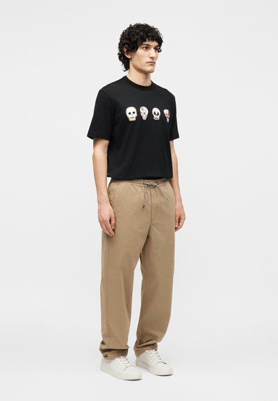 Νέος άνδρας με σγουρά μαλλιά, φορώντας μαύρο μπλουζάκι με τέσσερα σχέδια κρανίων, μπεζ παντελόνια με κορδόνι και λευκά αθλητικά παπούτσια, στέκεται μπροστά σε λευκό φόντο.