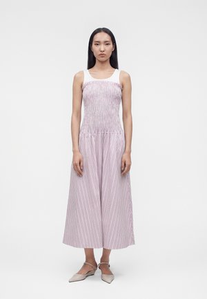 Femme debout portant une robe midi sans manches à rayures roses et blanches avec un corsage ajusté et une jupe large, ainsi que des ballerines beiges pointues.