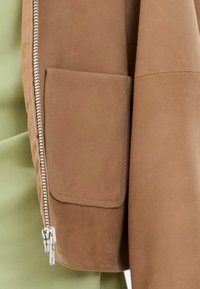 Κάμελ σουέτ τζάκετ με φερμουάρ μπροστά και ασημένια αξεσουάρ, τσέπη με διάτρητο σχέδιο στον ώμο και λεία υφή.