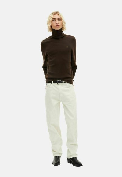 Brauner Rollkragenpullover, weiße gerade Jeans, schwarzer Gürtel und schwarze Knöchelstiefel. Das Model steht mit einem neutralen Ausdruck vor einem weißen Hintergrund.