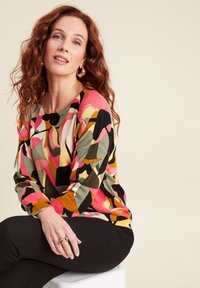 Multikleurige gebreide trui met een geometrisch patroon in roze, groen, zwart en gele tinten, gecombineerd met een zwarte broek.