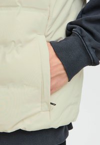 Svijetlosmeđi puffer prsluk s bočnom džepom, teksturirana tkanina i rebrasti tamnoplavi rukavi. Prsluk ima glatku, podstavljen završetak.