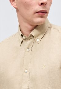 Κοντινό πλάνο ενός ατόμου που φοράει μπεζ λινό πουκάμισο με κουμπιά, με ένα μικρό κεντητό λογότυπο στο στήθος.