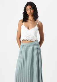Valge pitsist crop top, millel on konarate servad, koos helesinise volditud seelikuga. Kanga tekstuur näeb välja kerge ja voolav.