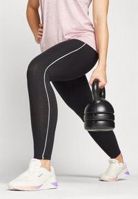 Sorte leggings med hvite sidestriper, kombinert med en lyserosa topp og hvite sneakers med lilla detaljer, som holder en svart kettlebell.