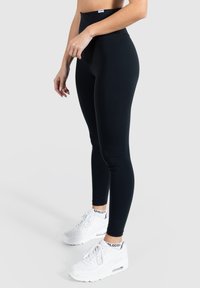 Mustat korkeavyötäröiset legginsit joustavasta kankaasta, jotka korostavat sulavaa istuvuutta. Yhdistettyinä valkoisiin lenkkareihin, joissa on näkyvää ilmapatjausta.