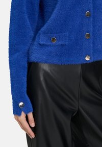 Blauwe fuzzy cardigan met zilveren knopen, gecombineerd met zwarte leren broeken, laat een close-up van de taille en de linkerhand zien.