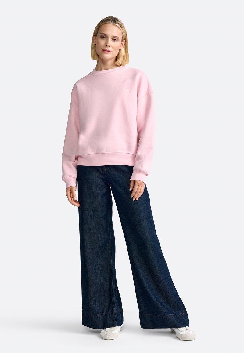 Felpa rosa chiaro con scollatura rotonda e polsini a coste, abbinata a pantaloni in denim scuro a gamba larga e sneakers bianche.