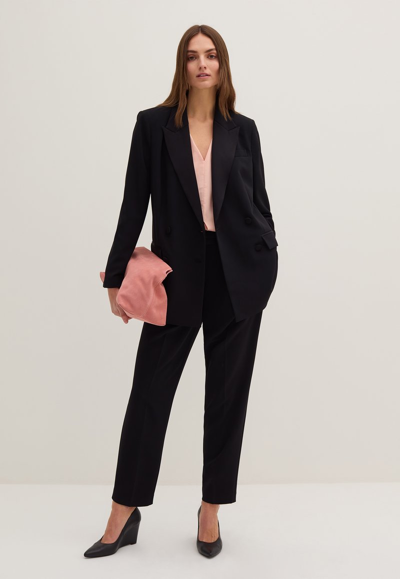 Schwarzer zweireihiger Blazer über einer hellrosa Bluse, kombiniert mit schwarzen Hosen und schwarzen Pumps. Hält eine weiche rosa Clutch.