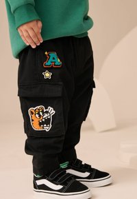 Fekete cargo nadrág hímzett foltokkal: színes "A" betű, csillag és meseállat tigris arc. Zöld pulcsi és fekete edzőcipő egészítik ki a megjelenést.