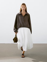 Giacca nera corta con bottoni e un grande collare, abbinata a una gonna bianca plissettata di lunghezza media. Sandali e una piccola borsa marrone completano il look.
