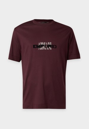 Un jeune homme se tient face à l’avant, portant un t-shirt logo brun foncé, un jean bleu clair et des baskets noires sur un fond clair uni.