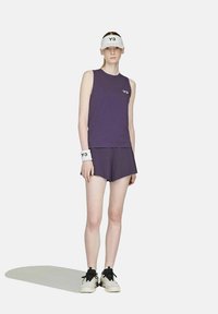 Eine weibliche Model trägt ein lila ärmelloses Top und Shorts, eine weiße Sonnenblende und ein Armband mit dem "Y-3"-Logo sowie schwarz-weiße Sneakers.