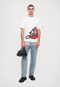 Camiseta blanca con un gráfico de un gato de dibujos animados en rojo y negro; jeans de mezclilla azul claro; bolso negro; zapatillas negras.