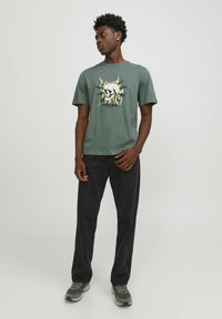 T-shirt en coton vert avec un design de crâne graphique et du texte, associé à un jean noir et des sneakers grises, offrant une coupe décontractée.