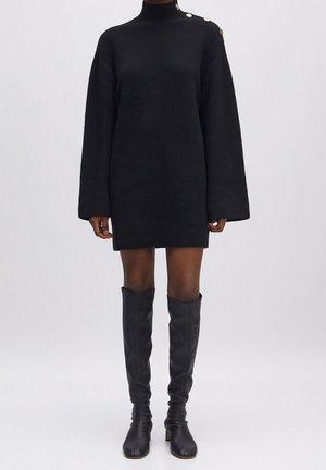 Schwarzes, geripptes Strickkleid mit goldenen Knöpfen an der Schulter, getragen mit kniehohen schwarzen Lederstiefeln an einem Model.