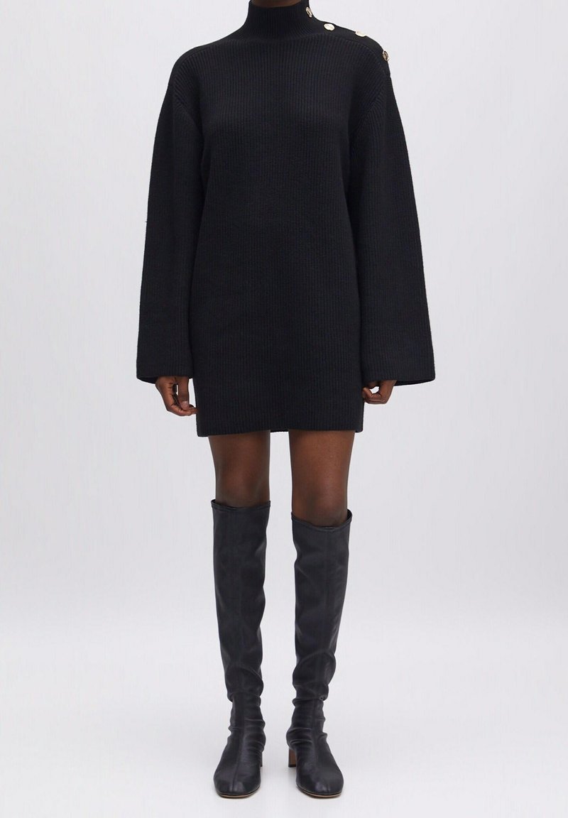 Robe pull noire côtelée avec boutons dorés sur l'épaule, portée avec des bottes en cuir noires montantes sur un mannequin.