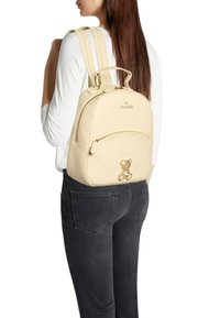 Zaino in pelle beige chiaro con forma rotonda, doppie bretelle e un orsetto decorativo sulla parte anteriore. Presenta una tasca frontale con cerniera.