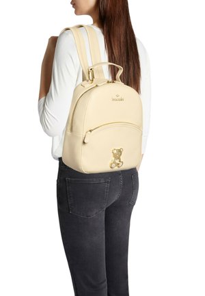 Zaino in pelle beige chiaro con forma rotonda, doppie bretelle e un orsetto decorativo sulla parte anteriore. Presenta una tasca frontale con cerniera.