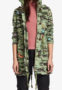 Grön jacka med camouflagemönster och huva, med fjärilsbroderi, cinchad midja och träknappar. Lättviktstyg och funktionella fickor.