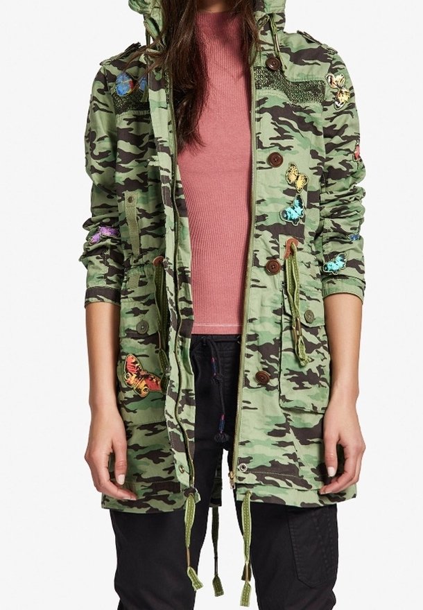 Grön jacka med camouflagemönster och huva, med fjärilsbroderi, cinchad midja och träknappar. Lättviktstyg och funktionella fickor.