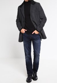 Uomo con cappotto grigio scuro, dolcevita nero, jeans blu e scarpe eleganti nere, in piedi con le mani in tasca su uno sfondo bianco.