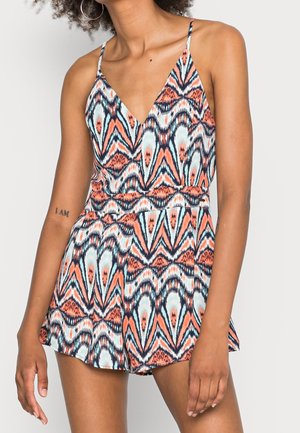 Femme portant une combinaison sans manches à col en V avec un motif géométrique multicolore en tons orange, noir, blanc et bleu.