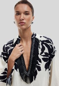 Blusa con estampado floral en crema y azul marino, con un profundo escote en V con borde de flecos negros y detalles en metal plateado. Tejido suave, corte relajado.