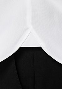 Witte shirtzoom met een driehoekige uitsnijding aan de achterkant, gecombineerd met een textuur zwart kledingstuk eronder. Gladde stof en strakke lijnen.