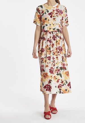 Femme portant une robe midi blanche avec des motifs floraux orange, jaune et bordeaux, ainsi que des sandales à talons ouvertes rouges.