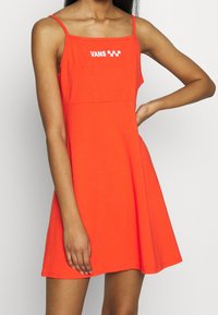 Robe sans manches orange avec un corsage ajusté et une jupe évasée. Présente un logo "VANS" blanc et un détail à motif à carreaux sur la poitrine.