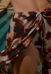 Zijdeachtige sjaal gemaakt van doorzichtig fabric met een multicolore bruin en beige luipaardprint, aan de zijkant geknoopt en elegant over het lichaam vallend.